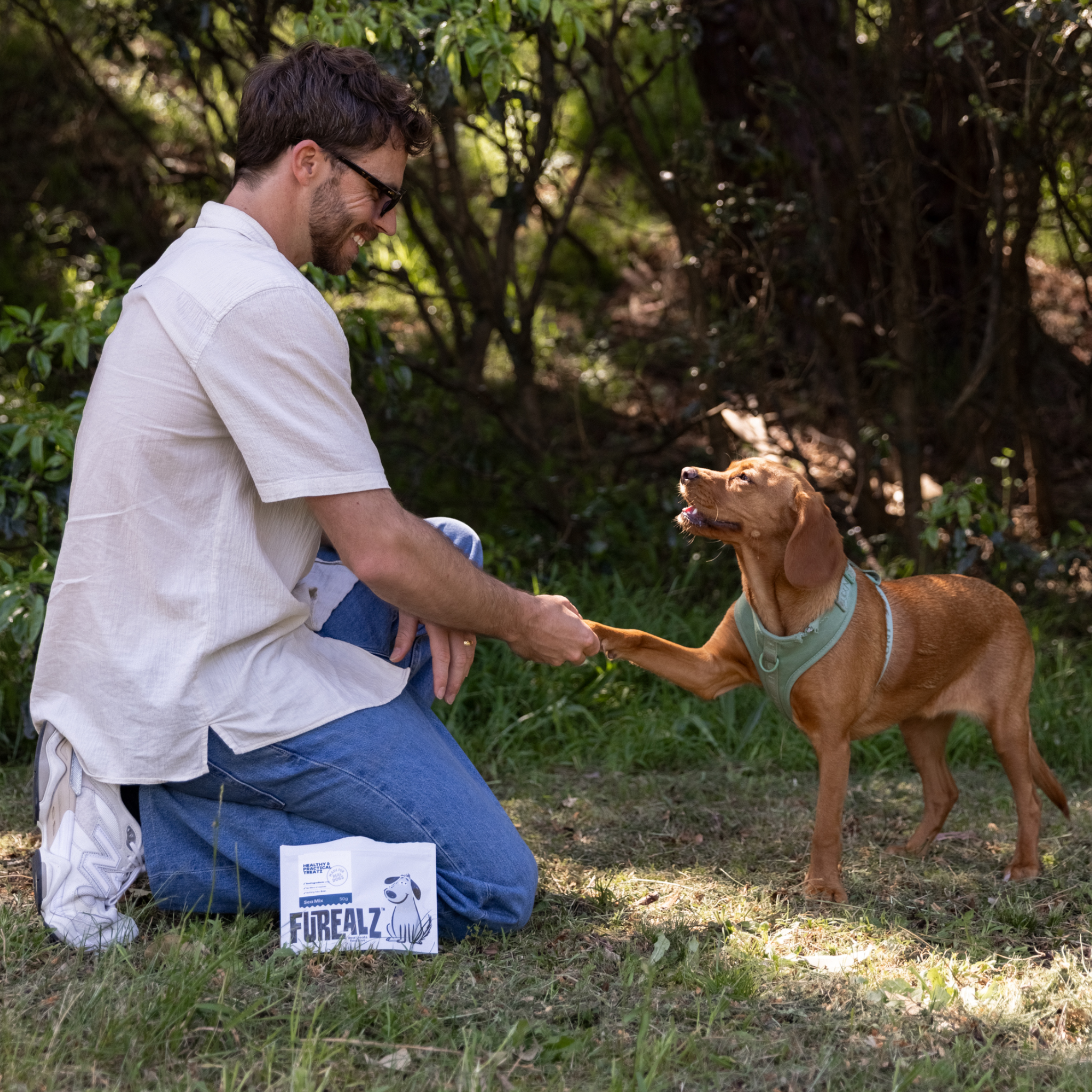 Man kneeling outdoors with a dog, with Furealz products.